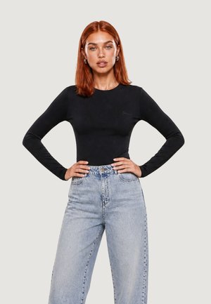 Femme aux cheveux roux portant un haut noir à manches longues et un jean bleu clair taille haute, debout les mains sur les hanches devant un fond uni.