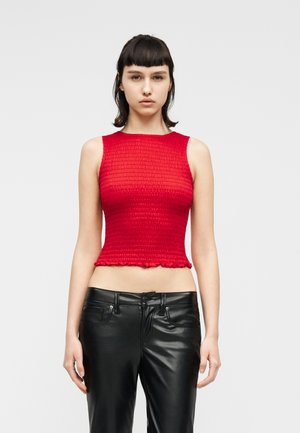 Femme aux cheveux courts et foncés portant un top court texturé rouge sans manches et un pantalon en cuir noir, debout devant un fond clair uni.