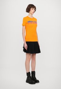 Oranžové bavlněné tričko s potiskem, v kombinaci s černou plisovanou sukní a černými bojovými botami. Jednoduchý, strukturovaný design.