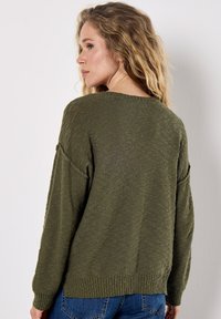 Pull en maille vert olive avec une coupe décontractée, des manches larges et un motif texturé. Bord-côte en bas et aux poignets. Porté avec un jean en denim.