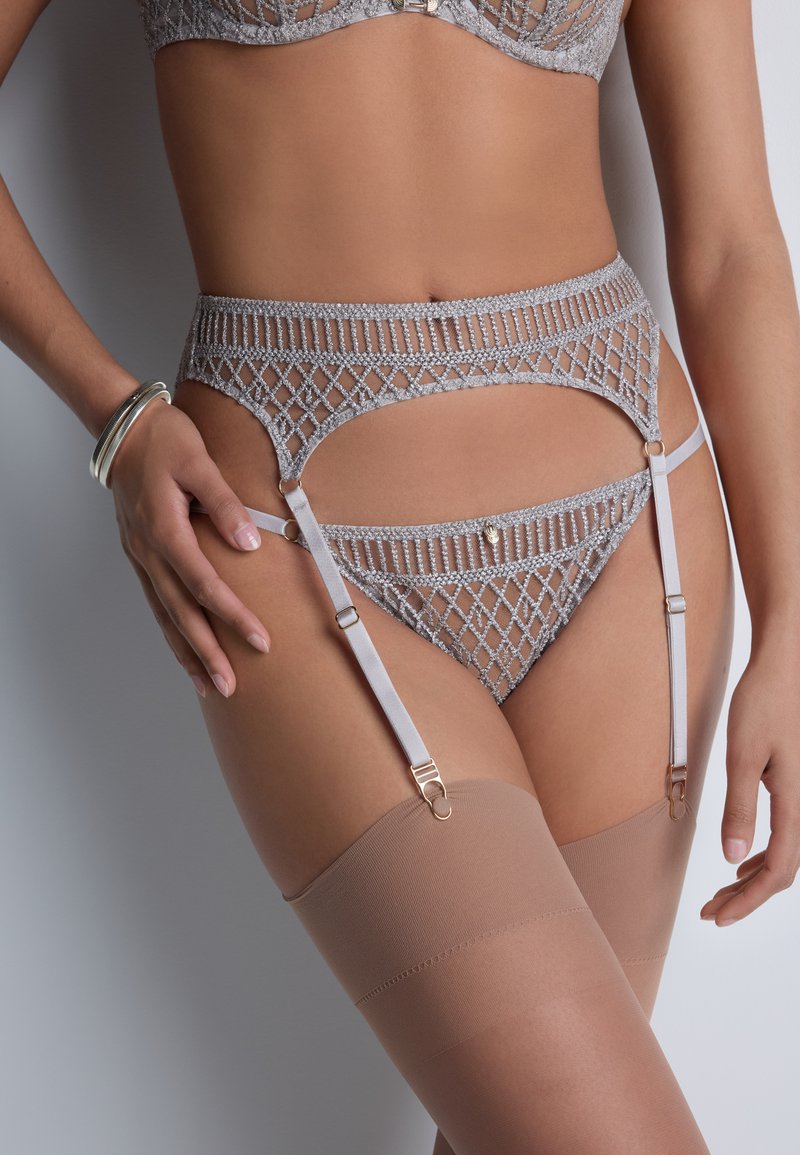 Ensemble de lingerie avec un soutien-gorge en dentelle argentée, une ceinture porte-jarretelles assortie, un string et des bas nude montants sur le haut des cuisses, posés sur la taille d'une femme.