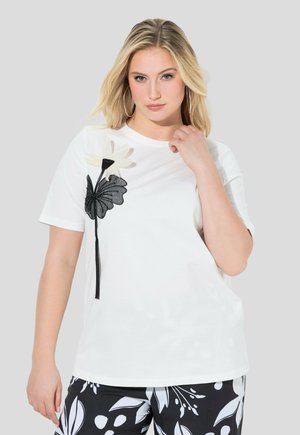 Blondynka w białej koszuli z krótkim rękawem z dużym czarno-białym wzorem kwiatowym, w parze z czarno-białymi spodniami w kwiaty.