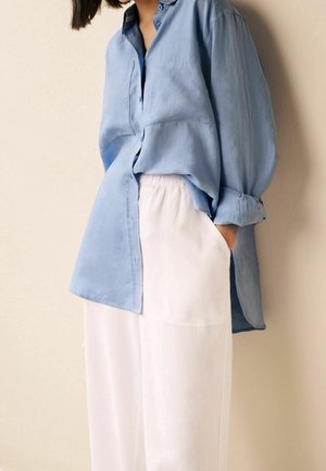 Personne portant une chemise ample bleu clair boutonnée avec les manches retroussées et un pantalon blanc taille haute, main dans la poche, debout contre un mur.