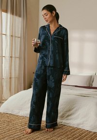 Navy floral pajama set in soft fabric. Features a button-up top with white piping, long sleeves, and matching loose pants with a subtle pattern.
