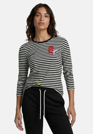 Frau trägt ein schwarz-weiß gestreiftes Langarmshirt mit Peanuts-Figuren und schwarze Hose mit weißen Kordeln.
