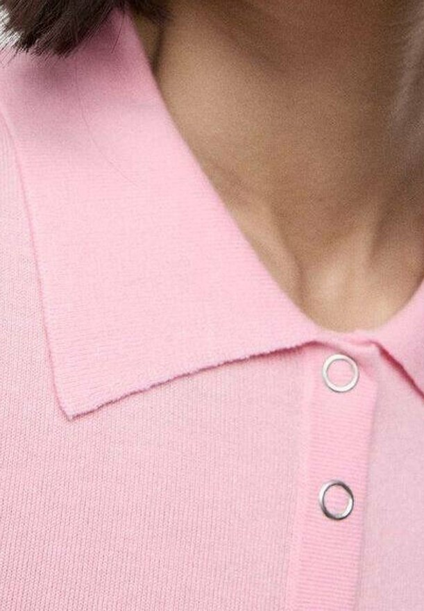 Gros plan d'une personne portant une chemise rose clair avec col et deux boutons-pression argentés visibles.