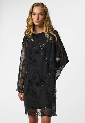 Mulher com cabelo loiro longo vestindo um vestido preto transparente com padrões florais e de lantejoulas, de pé contra um fundo claro simples.
