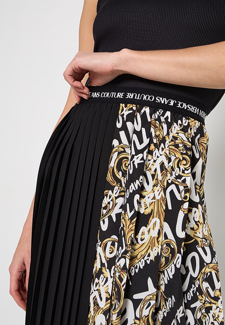 Frau, die die Taille eines schwarzen, gemusterten Plisseerocks mit dem Bund "Versace Jeans Couture" anpasst, trägt ein schwarzes ärmelloses Oberteil.