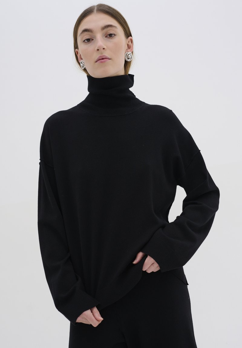 Melnā turtleneck džemperis no mīksta materiāla, oversized piegriezums, ar garām piedurknēm un ribotām malām. Iespējas iekļauj augstu apkakli un sānu šķēlumas.