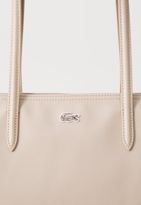 Sac fourre-tout beige avec un tissu texturé, deux longues anses et un petit logo crocodile brodé à l'avant.