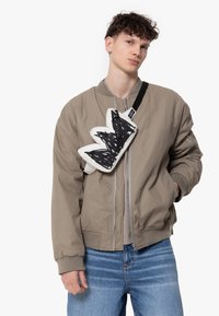Beige bomberjack met geribde manchetten, gedragen over een grijs shirt. Zwart-witte tas in abstracte vorm, kruislinks gedragen. Blauwe jeans.