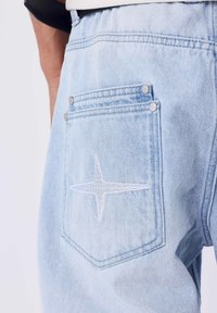 Pantaloni in denim azzurro chiaro con una tasca posteriore decorata da un ricamo di una stella bianca e due bottoni a pressione argentati. Texture liscia e design classico.