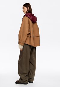 Brązowa, oversize'owa kurtka z burgundowym, puchowym kapturem, z warstwowym tyłem i białymi futrzanymi akcentami na rękawach. Luźne, oliwkowozielone spodnie.