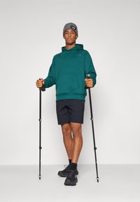 Felpa teal con tasca frontale e logo viola, abbinata a pantaloni corti neri e scarpe da trekking nere. Beanie grigio e bastoncini da trekking inclusi.