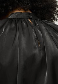 Blusa nera in satin con collo alto, retro plissettato e apertura a chiave fissata con un bottone sul collo. Texture liscia.