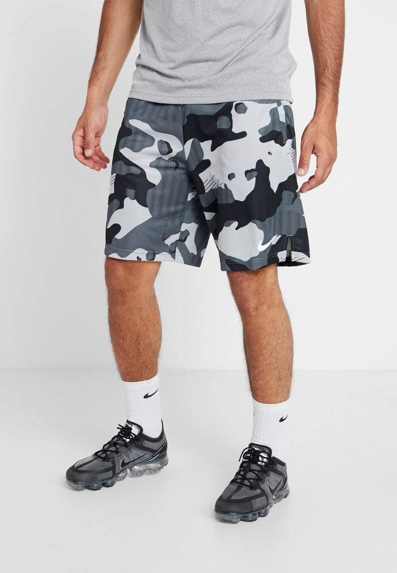 Homme portant un short de sport camouflage gris et noir, un t-shirt gris, des chaussettes blanches Nike et des baskets noires, debout sur un sol blanc.
