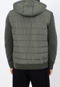 Gilet rembourré vert avec une couche de tricot texturée en dessous, doté d'une capuche, d'un design matelassé horizontal et d'un ourlet côtelé, adapté pour une tenue décontractée.