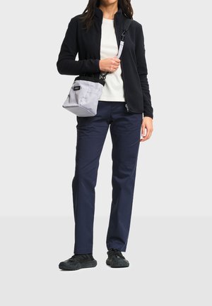 Woman wearing a black zip-up jacket, white shirt, navy pants, black sneakers, and carrying a small light gray crossbody bag.