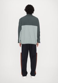 Marmoreret to-farvet jakke med grønne og turkise sektioner, elastisk talje, afslappet pasform, parret med navy sweatpants med brune og hvide accenter.