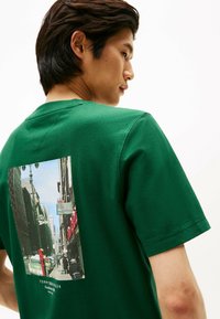 Grøn bomulds T-shirt med et fototryk på ryggen. Trykket viser en urban gadescene med bygninger, biler og et gadeskilt.