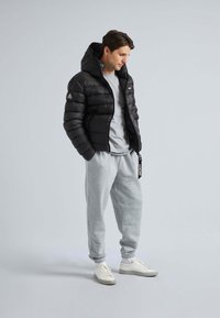 Sort pufferjakke med hætte, lynlås og sidelommer. Bæres over en grå t-shirt og lysegrå sweatpants med snor. Hvide sneakers.
