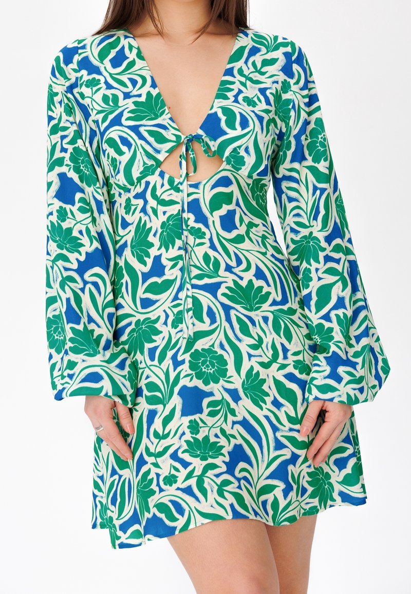 Femme portant une robe à manches longues à motifs floraux bleus, verts et blancs, avec une ouverture en forme de goutte d'eau nouée au niveau de la poitrine.