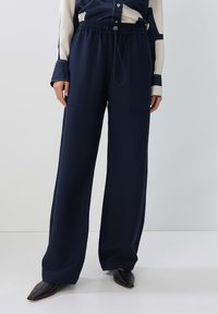 Navy wide-leg trousers with an elastic waistband, two front pockets, and a smooth texture. Paired with a long-sleeve blouse and pointed shoes.