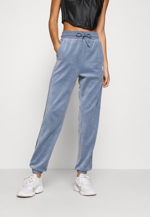Personne portant un pantalon de jogging en velours bleu avec une taille à cordon et des baskets blanches, debout devant un fond clair uni.