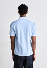 Persoon staand met zichtbare rug, gekleed in een lichtblauw polohemd met korte mouwen en een donkerblauwe broek tegen een effen achtergrond.