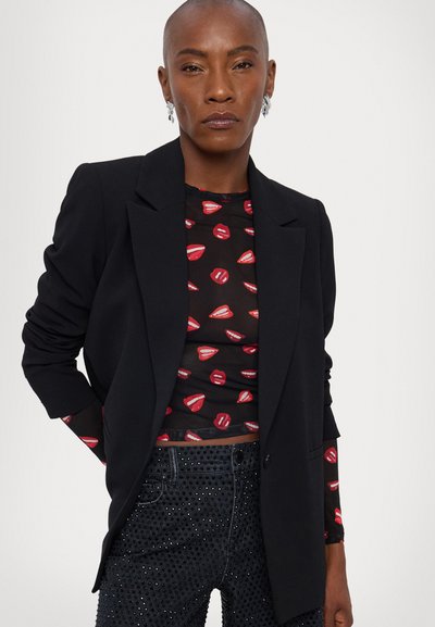 Black blazer over a sheer top featuring red lip patterns, paired with dark beaded high-waisted jeans. Sleek design and fitted silhouette.
