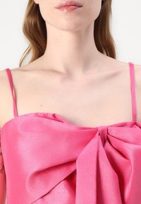 Rožinė atlasinė palaidinė su plonais petnešėlėmis, su surištos priekinės dalies dizainu ir lygia tekstūra. Audinys turi švelnų blizgesį ir elegantiškai krinta.