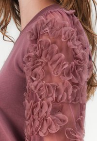 Top a maniche lunghe in mauve con applicazioni floreali a strati e texture sulle maniche in rete trasparente. Tessuto morbido con scollatura a costine.
