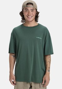 Jonge man die glimlacht in een groen oversized t-shirt, beige shorts, een beige achterstevoren gedragen pet, en zichtbare tatoeages op beide onderarmen.