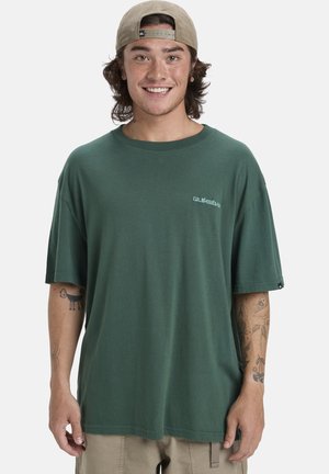 Young man smiling wearing a green oversized t-shirt, beige shorts, beige backward cap, and visible tattoos on both forearms.