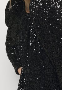 Czarna, cekinowa bluzka z długim rękawem, z bufkami na ramionach, teksturowana tkanina, ręka w kieszeni, oraz widoczne długie, złote kolczyki.