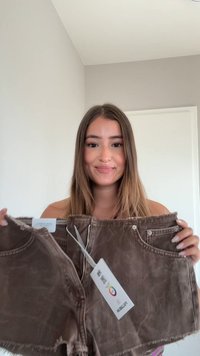 Shorts di jeans marroni con orlo sfrangiato, zip frontale e tasche. Etichetta attaccata che mostra il marchio e le informazioni per la cura.