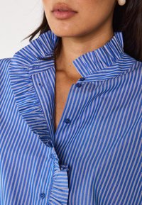 Camicia a righe blu e bianche con colletto arricciato, chiusure a bottoni e texture liscia, che mette in risalto sottili linee verticali e un design su misura.