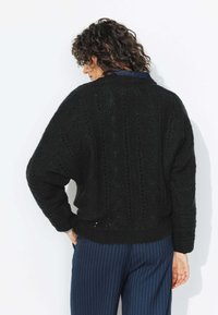 Pull en maille noir avec des motifs texturés, coupe oversized et ourlet à côtes. Porté avec un pantalon bleu à rayures.
