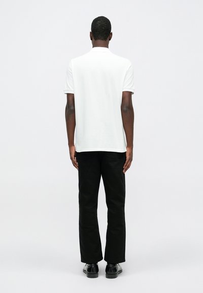 Tricou polo alb cu mânecă scurtă, purtat cu pantaloni negri și pantofi negri. Design minimalist, fără modele sau logo-uri vizibile.