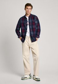 Camicia a quadri botton-up blu navy e rossa, indossata sopra una t-shirt bianca con grafica. Pantaloni beige chiaro e sneakers bianco e nere con accenti verdi.
