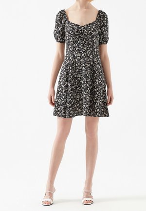 Femme portant une robe courte noire à fleurs avec des manches bouffantes et un décolleté carré, assortie à des sandales blanches à lanières, debout sur fond blanc.