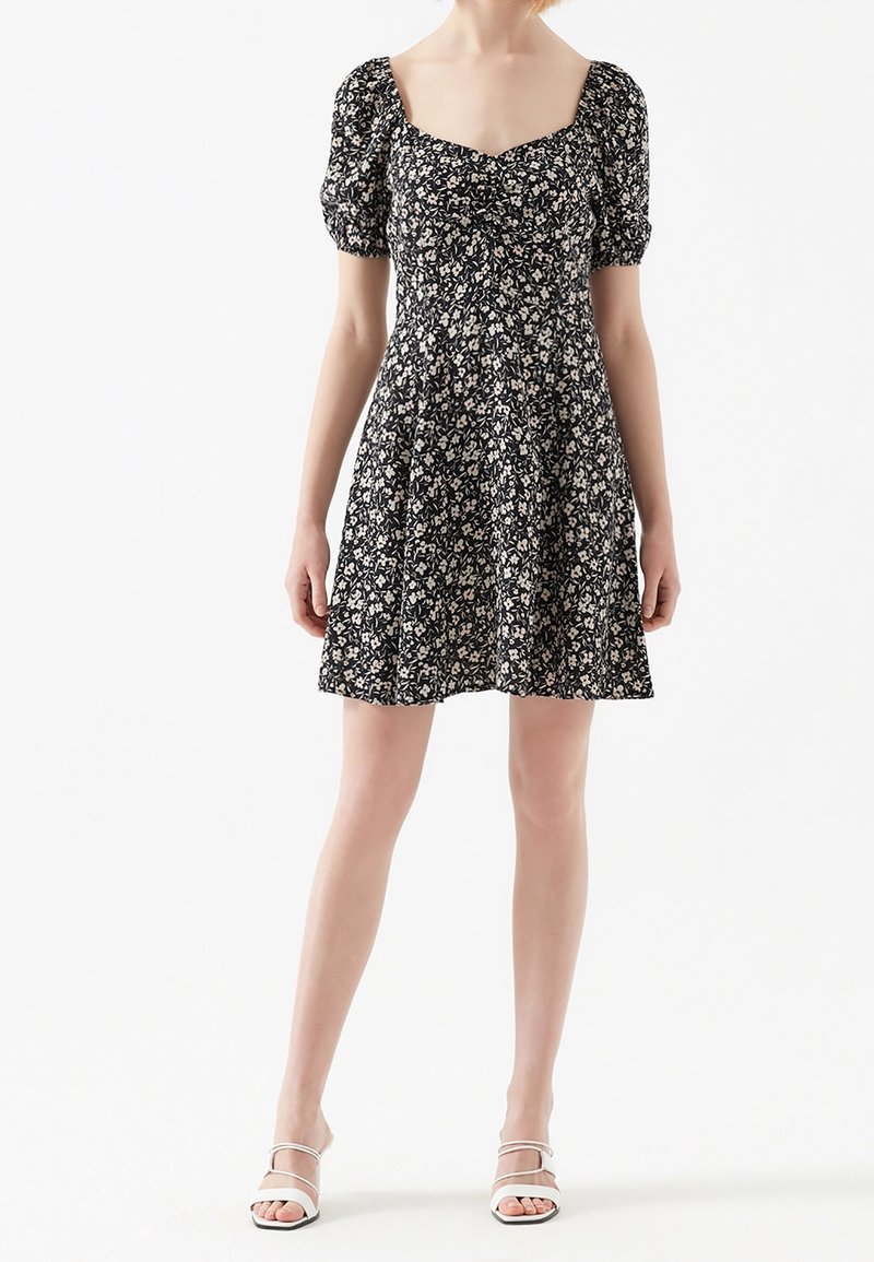 Femme portant une robe courte noire à fleurs avec des manches bouffantes et un décolleté carré, assortie à des sandales blanches à lanières, debout sur fond blanc.
