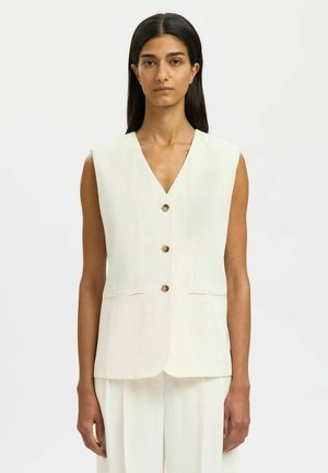 Frau mit langem dunklem Haar trägt ärmellose weiße Weste mit Knöpfen und passende weite Hosen, steht vor einfachem hellem Hintergrund.