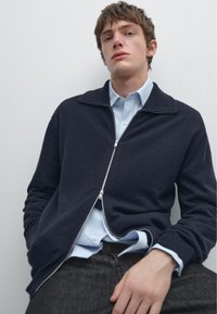 Námořnický zipový svetr vyrobený z texturované tkaniny, se vysokým límcem a stříbrným zipem. Nesený přes světle modrou košili s detaily knoflíků.