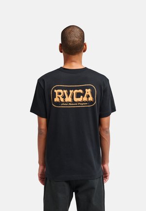 Uomo che indossa una t-shirt nera con un grande logo beige RVCA e la scritta "Artist Network Program" sulla schiena, in piedi davanti a uno sfondo semplice.