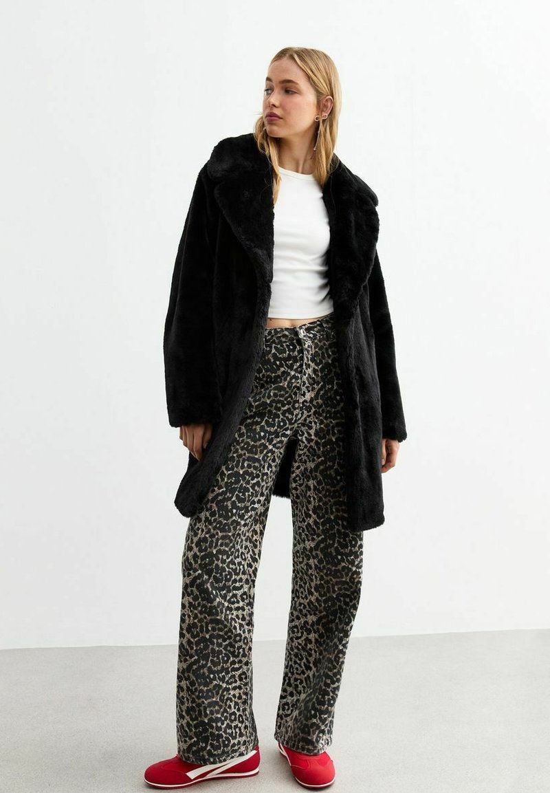 Abrigo de piel sintética negra con un amplio cuello, llevado sobre pantalones de pierna ancha con estampado de leopardo, combinado con zapatos rojos de estilo slip-on. Textura suave, diseño casual.
