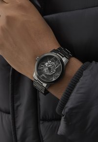 Zwart metalen horloge met een gestructureerde zwarte marmeren wijzerplaat met een reliëfontwerp. Zilveren wijzers en een schakelband maken de uitstraling compleet.