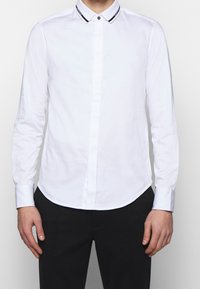 Vit långärmad skjorta med krage kantad i svart och knappstängning, med en mjuk textur och rundad nederkant.