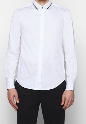 Camicia bianca a maniche lunghe con colletto a righe nere e chiusura con bottoni, dotata di una texture liscia e orlo arrotondato.