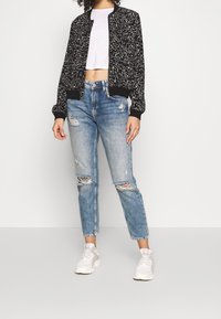 Svart bomberjacka med vitt abstrakt mönster, kombinerad med ljusblå slitna jeans. Vita sneakers fulländar outfiten.